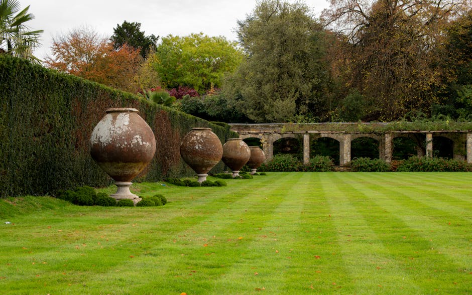 A well-maintained garden lawn with evenly cut, vibrant green grass extending across the foreground, bordered by a neatly trimmed hedge on the left side. Along the hedge, there are five large, weathere