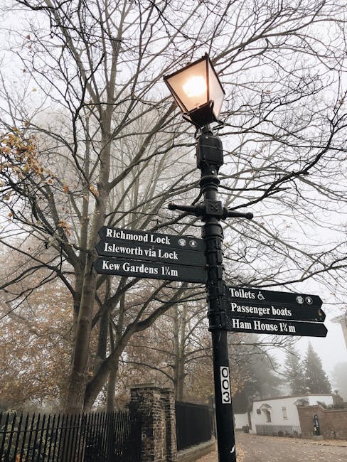 A black vintage-style street lamp emitting warm light is mounted on a tall black lamppost situated in a garden area. The lamppost features several black directional signs with white text pointing towa